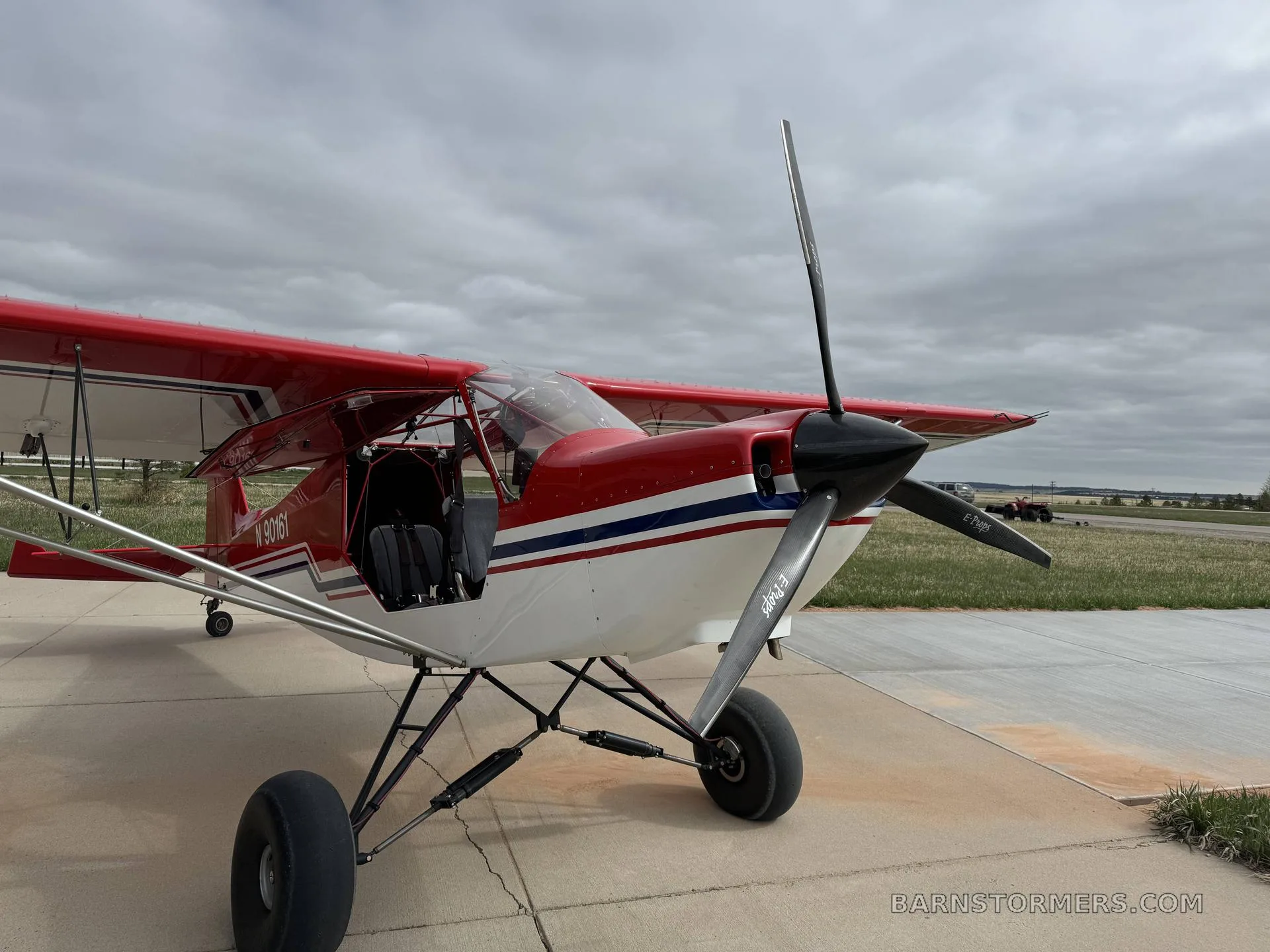 A red and white single-engine STOL aircraft with large tires parked on a concrete pad under a cloudy sky.