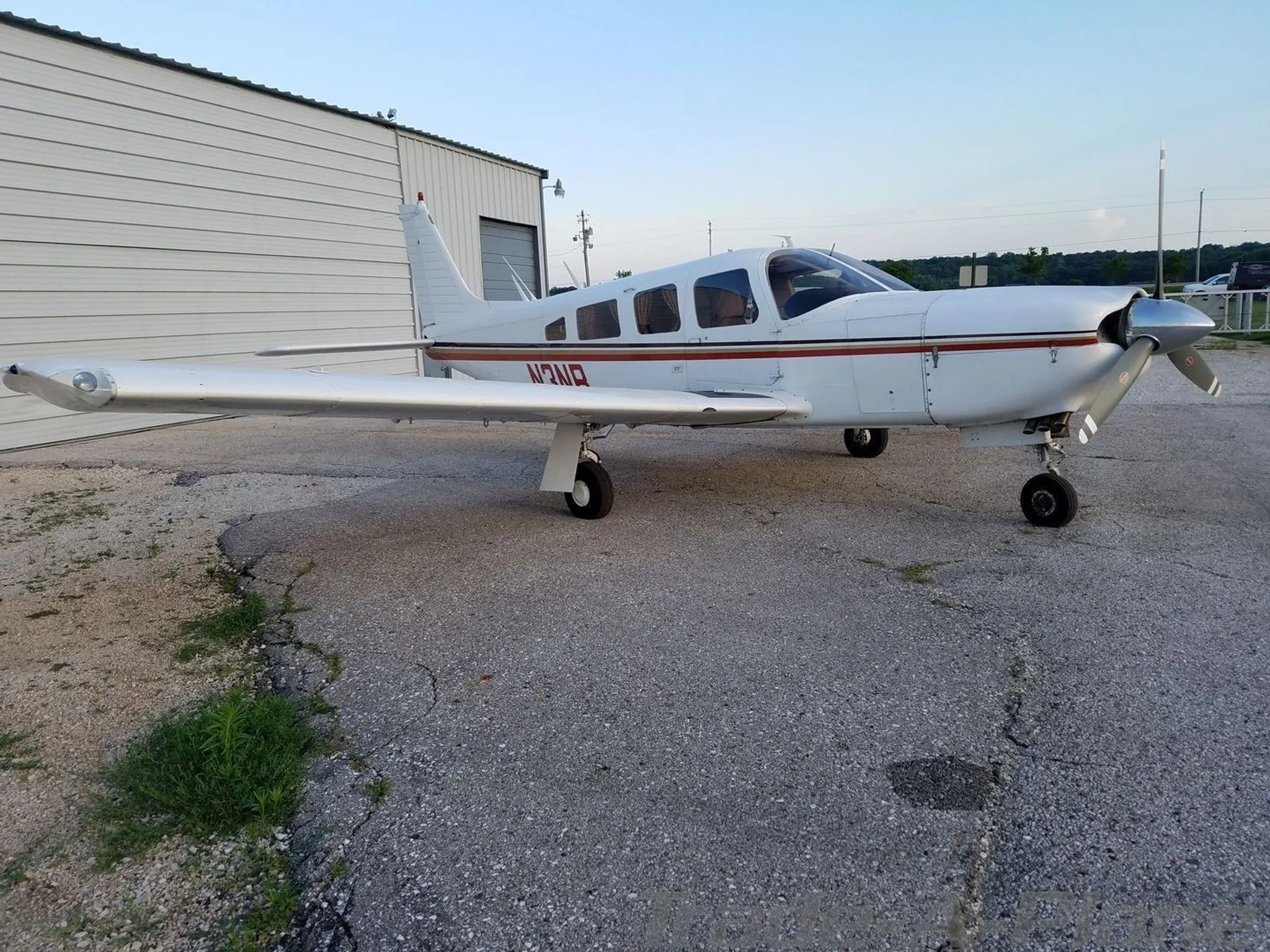 A white single-engine Piper aircraft with red and brown stripes, registration N3NB, parked on an asphalt apron next to a hangar.