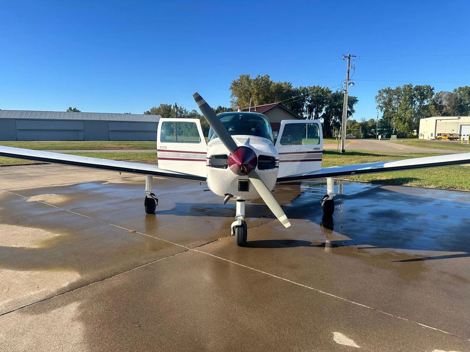 Front view of a white and maroon Beechcraft Sundowner 180 on a wet tarmac, with doors open.