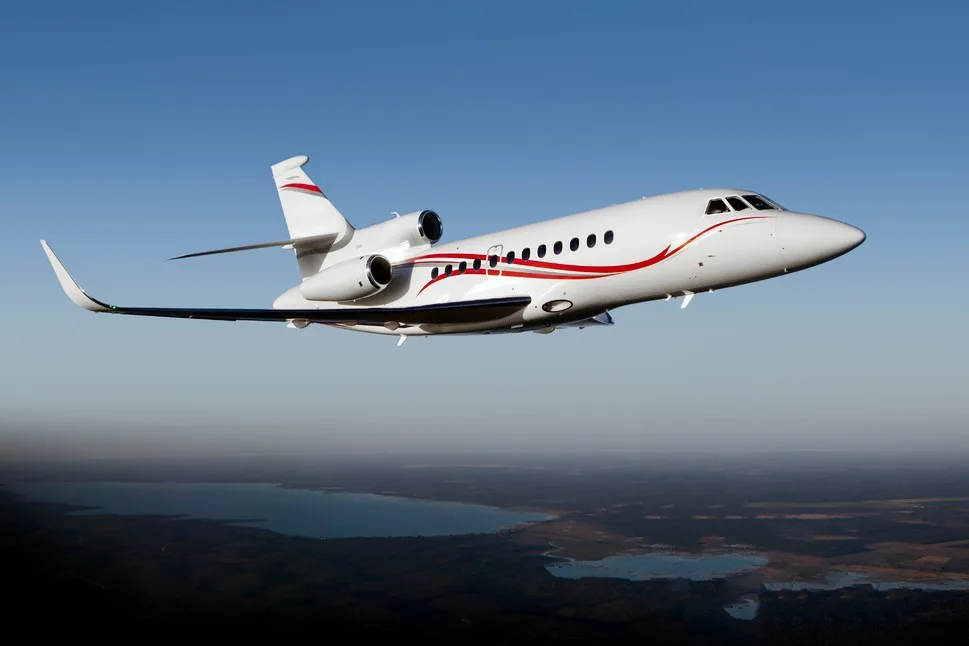 A white business jet with red stripes flies over a landscape with lakes under a clear blue sky.