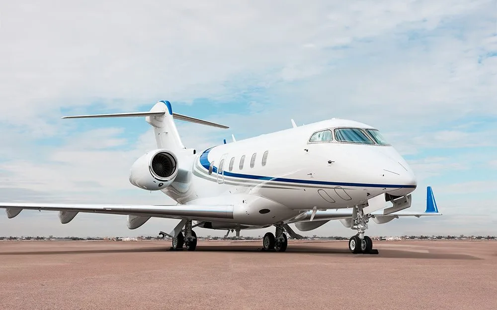 A white private jet with blue accent stripes is parked on an airfield under a partly cloudy sky.