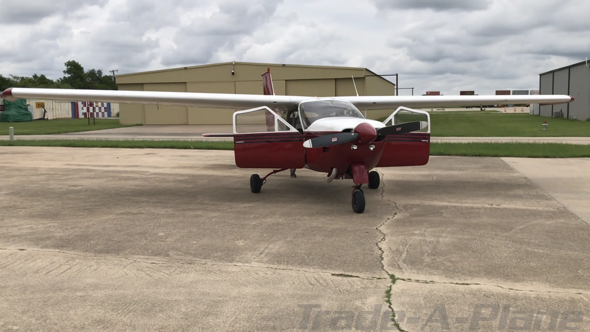 A red and white high-wing single-engine aircraft, N33297, with its doors open, parked on an asphalt ramp in front of hangars under a cloudy sky.