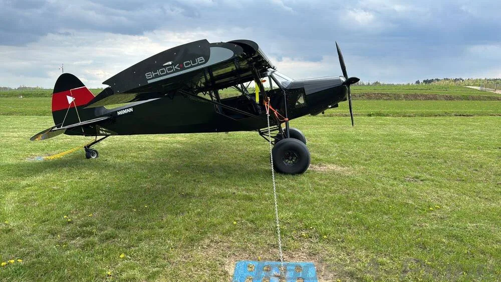 A black Zlin Shock Cub aircraft, registration N696NN, parked and tied down on a green grassy field under a cloudy sky.