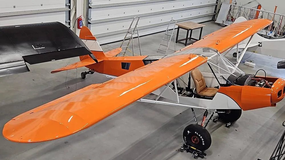 An overhead view of an orange and white high-wing aircraft with its engine cowling open, parked in a hangar.