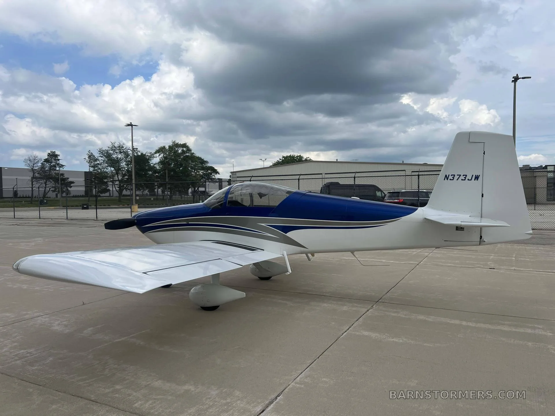 A white and blue single-engine aircraft, N373JW, with a bubble canopy, parked on a concrete tarmac with a fence and trees in the background under a cloudy sky.