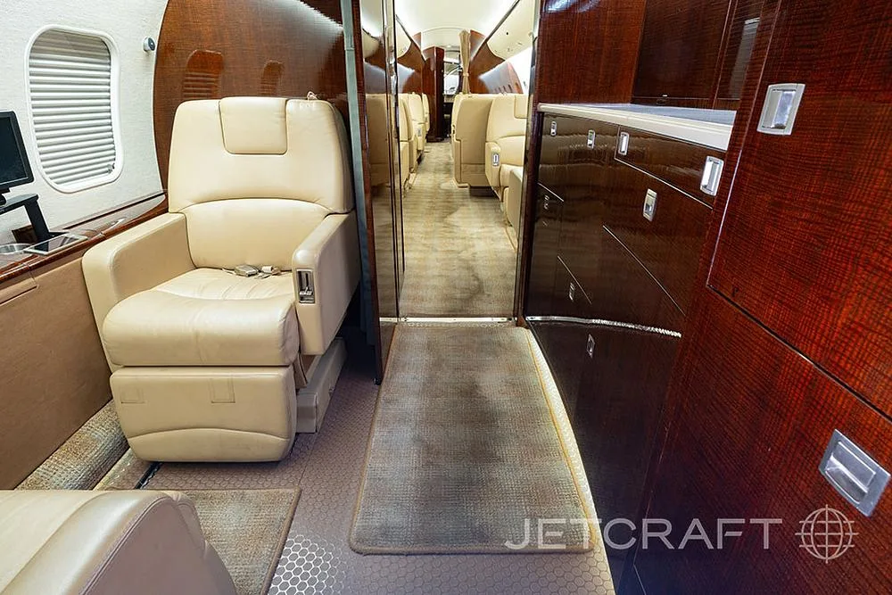 A view down the aisle of the private jet cabin, showing beige seats and dark wood cabinetry.
