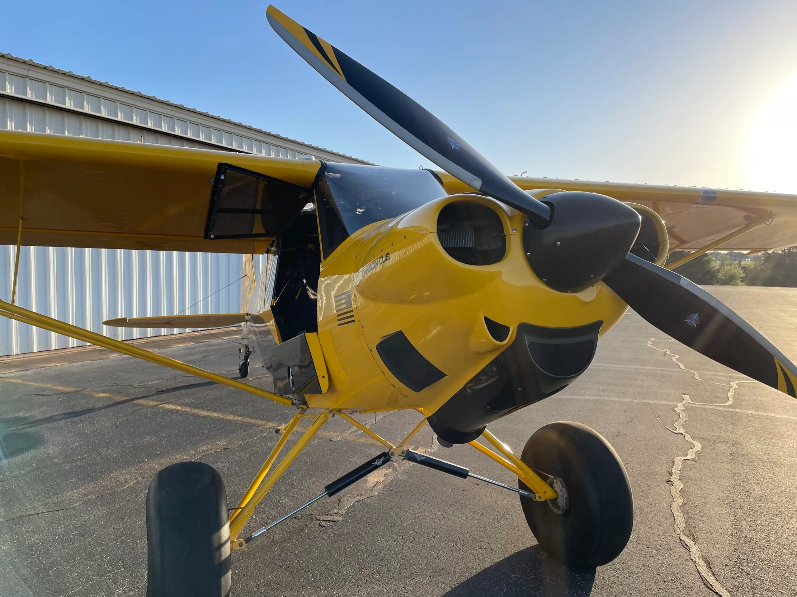 A bright yellow Carbon Cub aircraft with black accents and a propeller, parked on an asphalt apron with a hangar in the background.