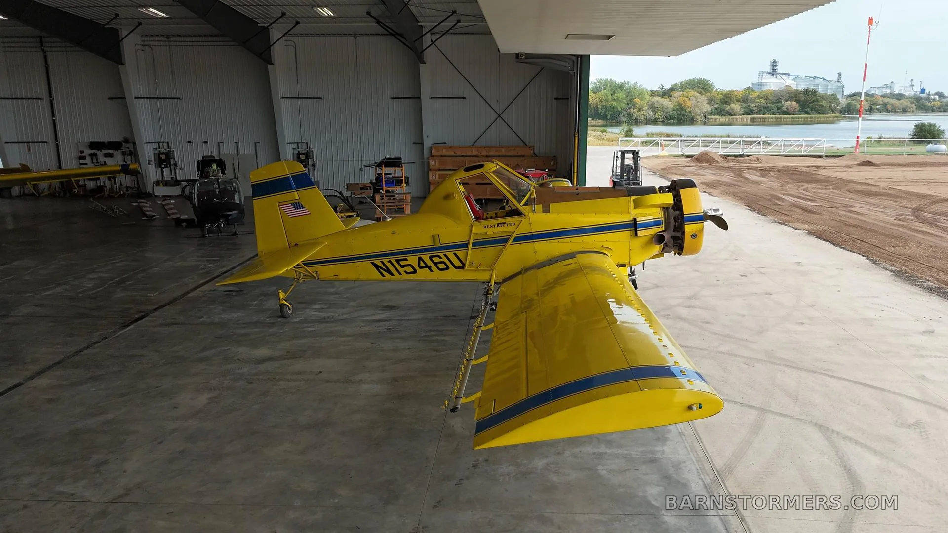 A yellow agricultural aircraft with blue stripes (N1546U) parked inside a hangar, with a view of a lake and industrial buildings outside.