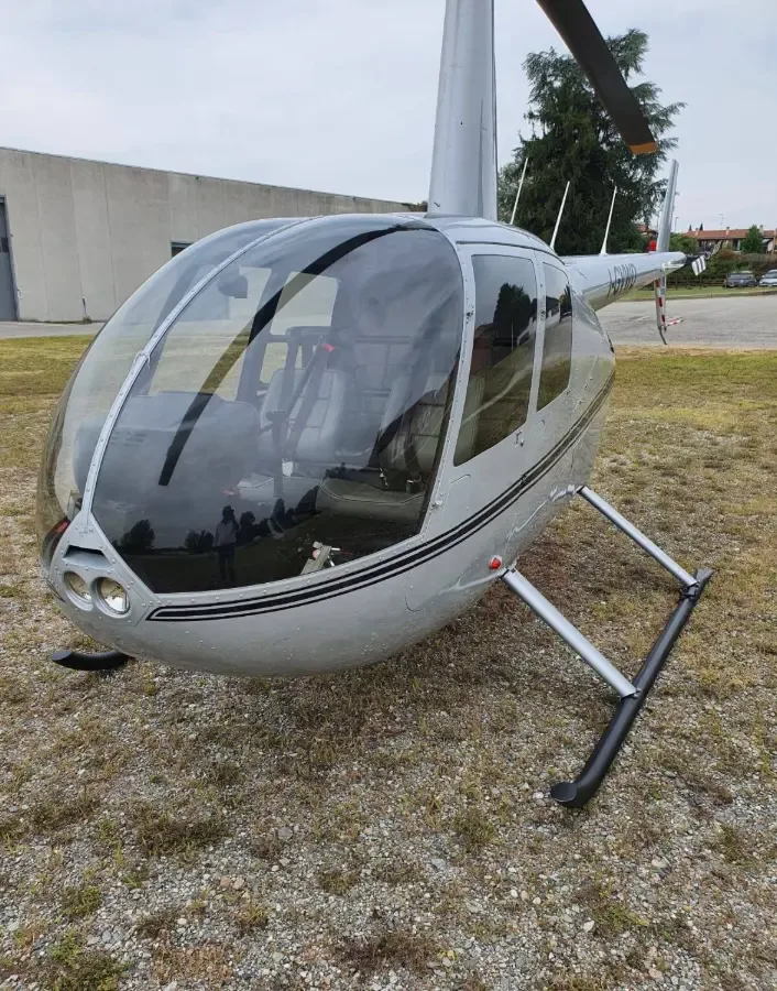 A close-up front-side view of a silver Robinson R44 helicopter on a gravel patch.