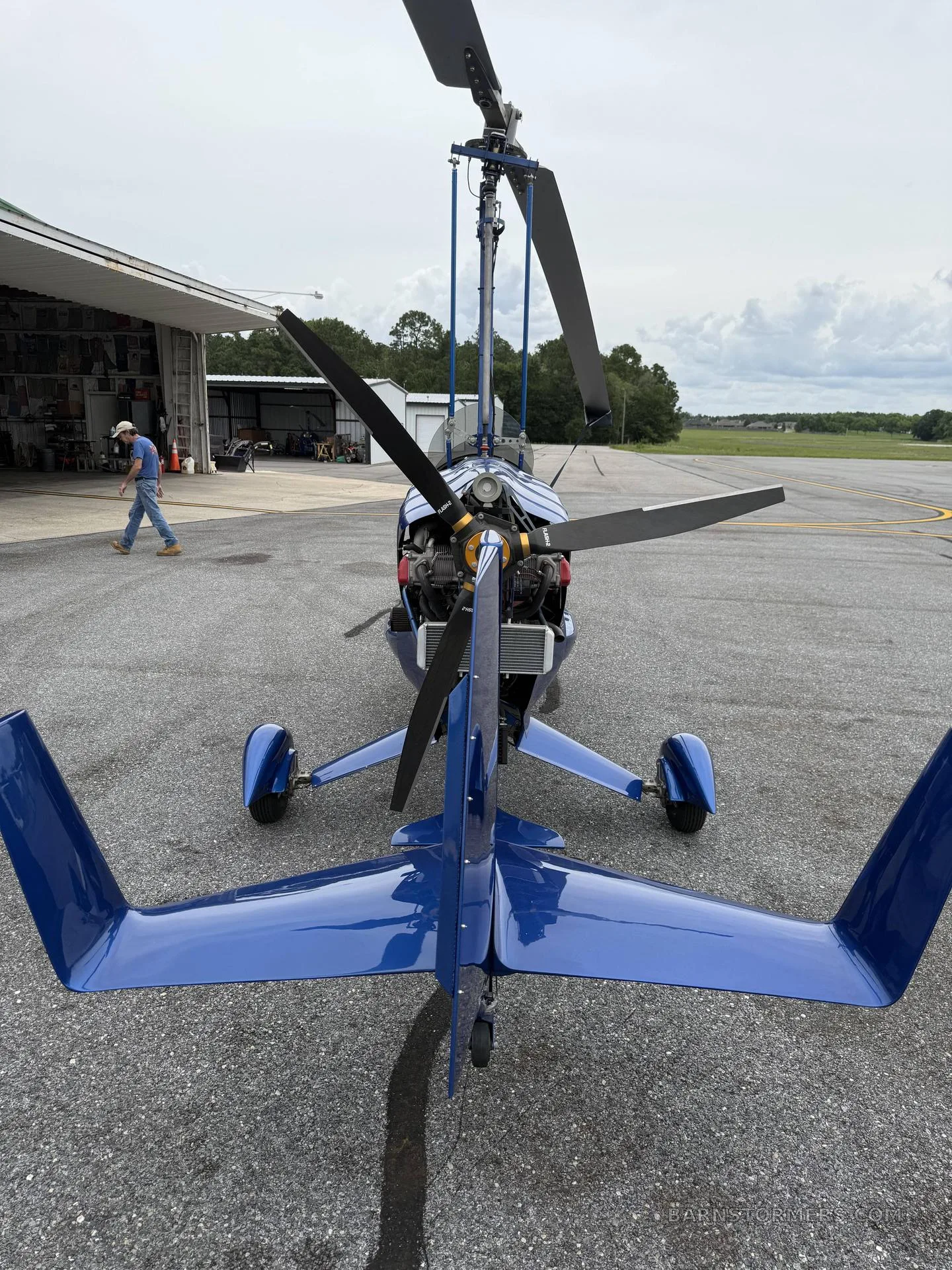 A blue autogyro is parked on an asphalt tarmac, viewed from the rear, with a person walking near a hangar in the background.