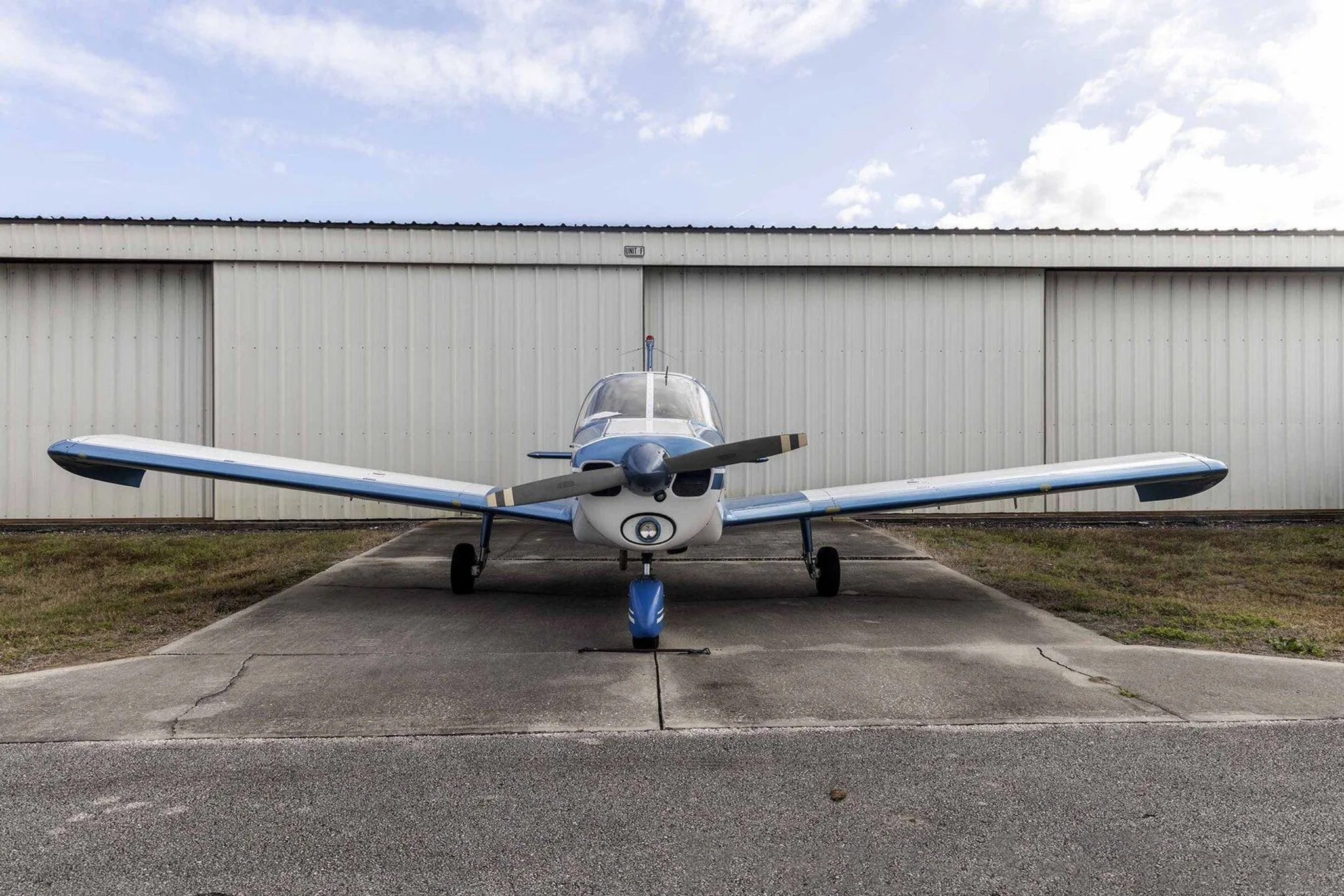 A wide front view of the blue and white Piper Cherokee N586 parked on an asphalt ramp in front of a hangar.