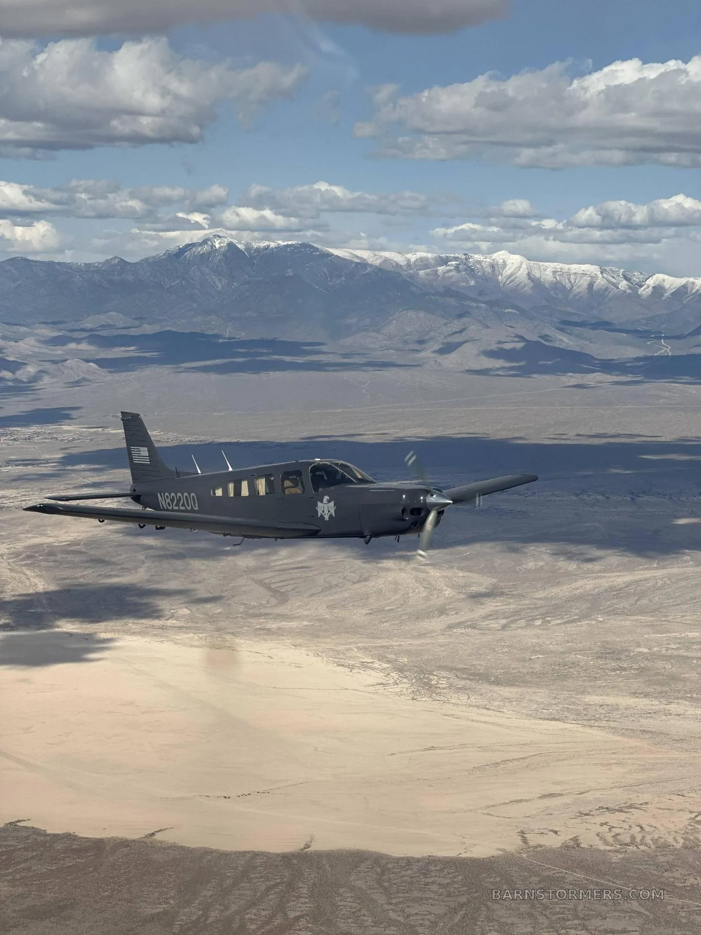 A gray single-engine propeller aircraft, N8220Q, flying over a desert landscape with snow-capped mountains in the background under a partly cloudy sky.