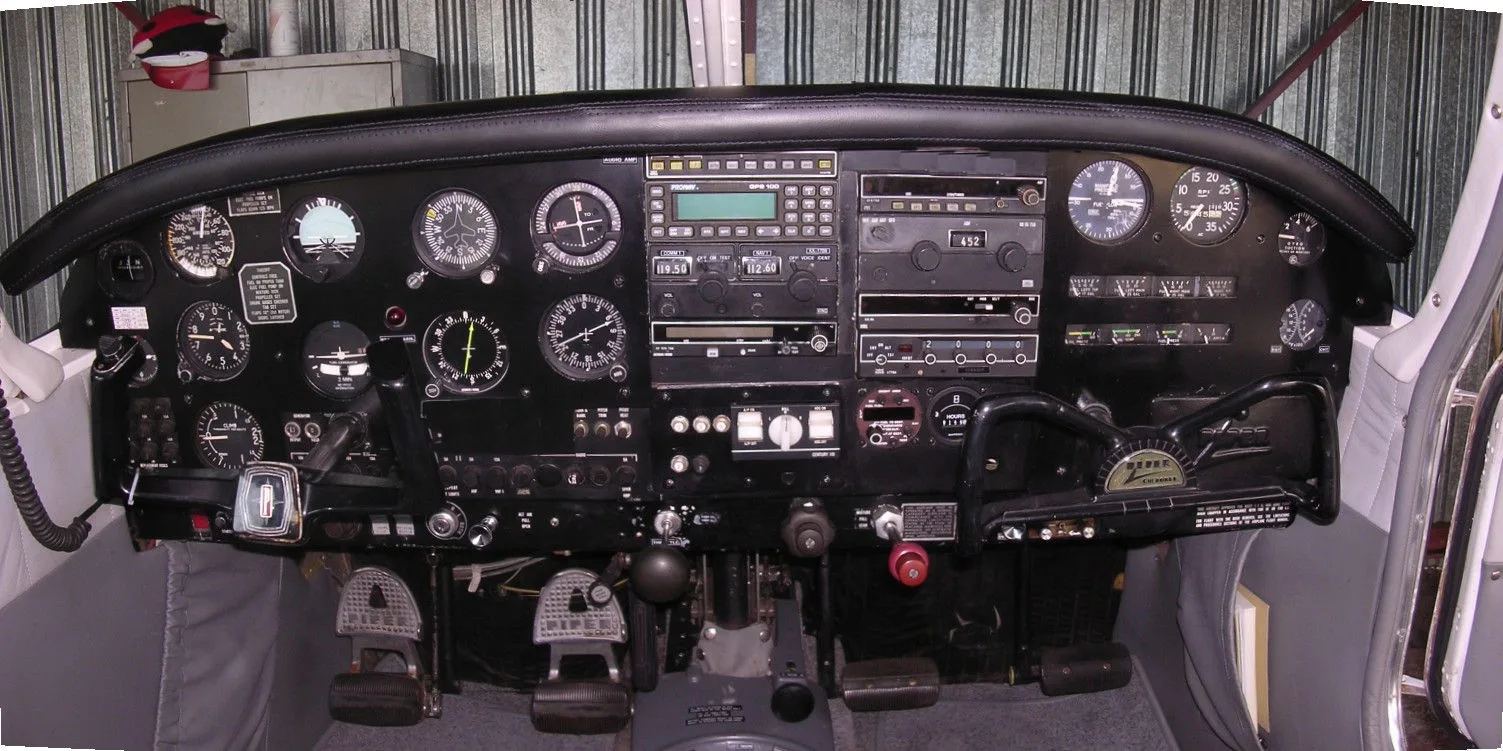 A detailed view of an aircraft cockpit with a black instrument panel, various gauges, and avionics.