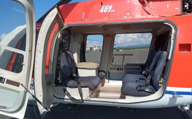 The open cabin door of a red and white helicopter reveals two rows of dark grey seats and a view of the airfield outside.
