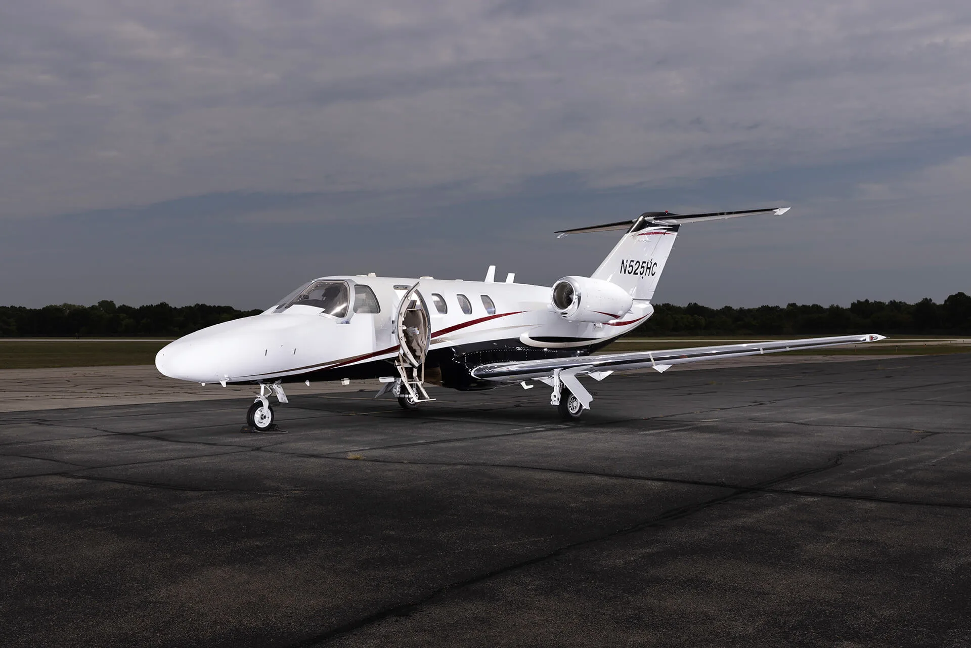 A white private jet with black and red stripes and an open door is parked on an asphalt tarmac under a cloudy sky.