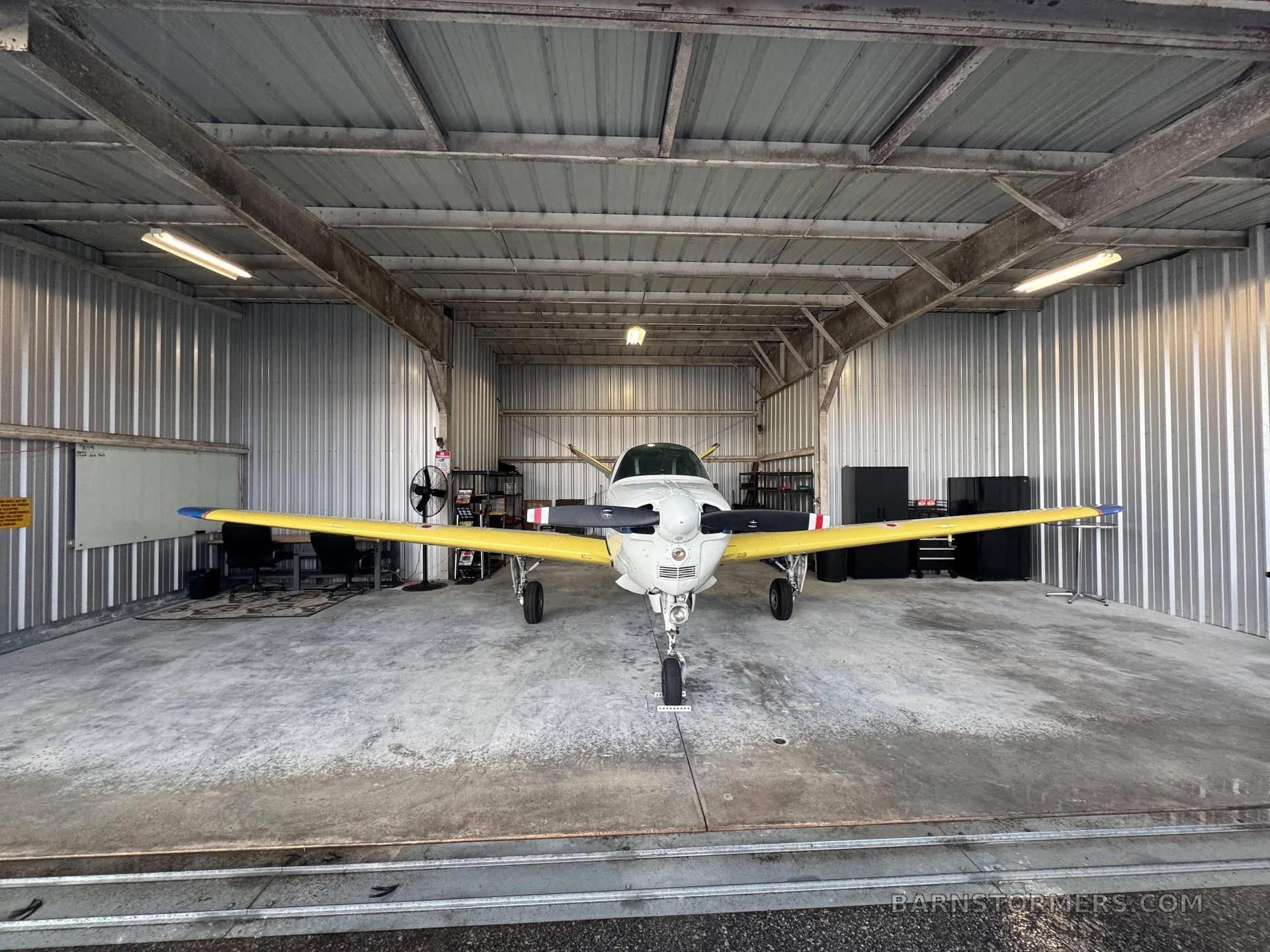 A white and yellow single-engine aircraft with a propeller, viewed head-on inside a metal hangar.