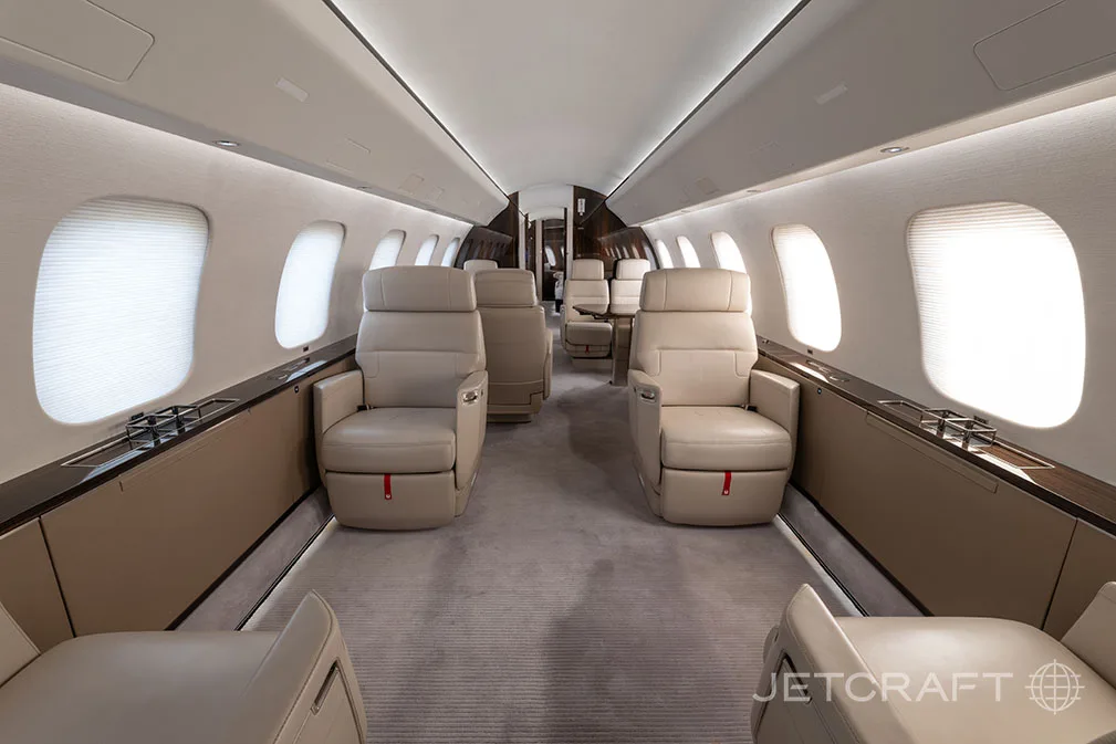 The luxurious interior of a business jet cabin features light beige leather seats and a grey carpet, viewed from the front looking towards the rear.