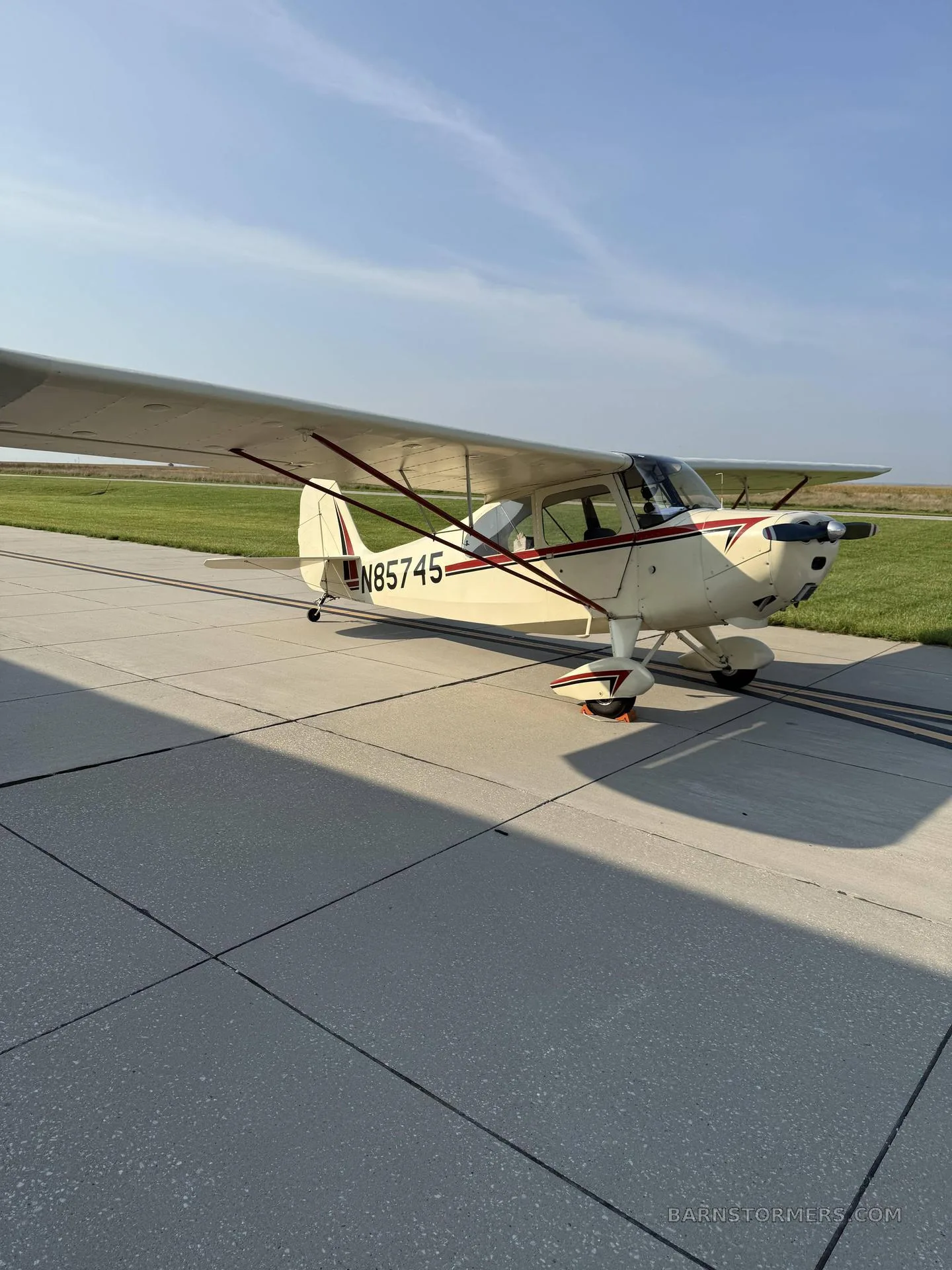 A cream-colored, high-wing taildragger aircraft with red and black stripes, registration N85745, parked on a concrete tarmac under a clear sky.