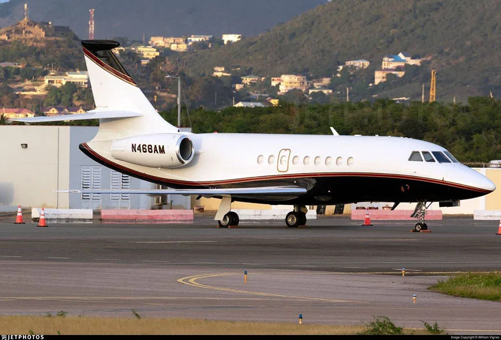 A white and black Dassault Falcon 900EX, tail number N468AM, parked on an airport tarmac with hills and buildings in the background.