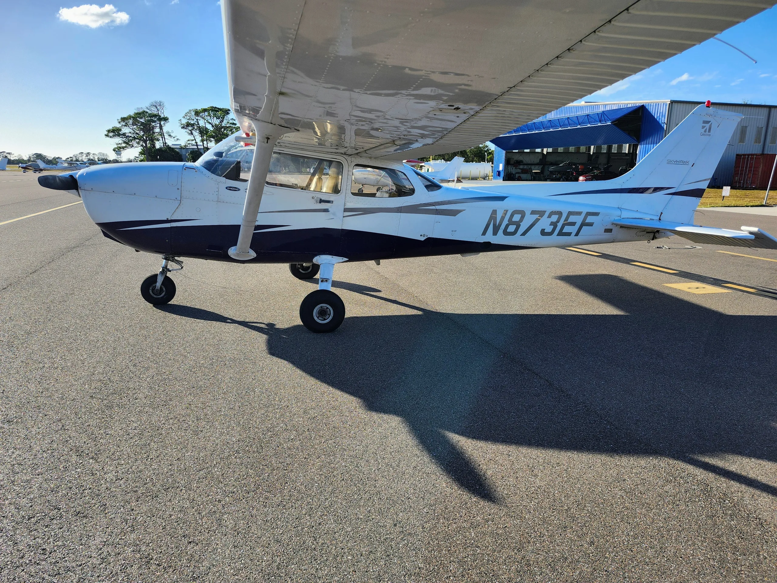 A white and blue Cessna 172, registration N873EF, parked on an airport tarmac on a sunny day.