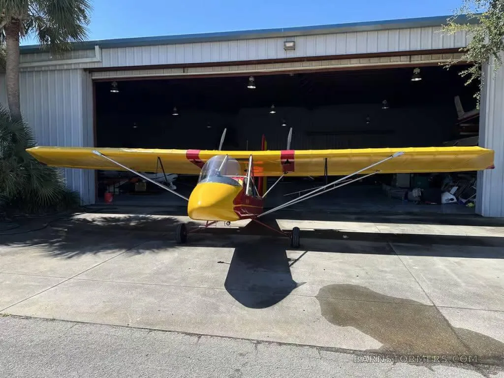 A red and yellow Air-Cam ultralight aircraft is parked head-on outside a hangar.