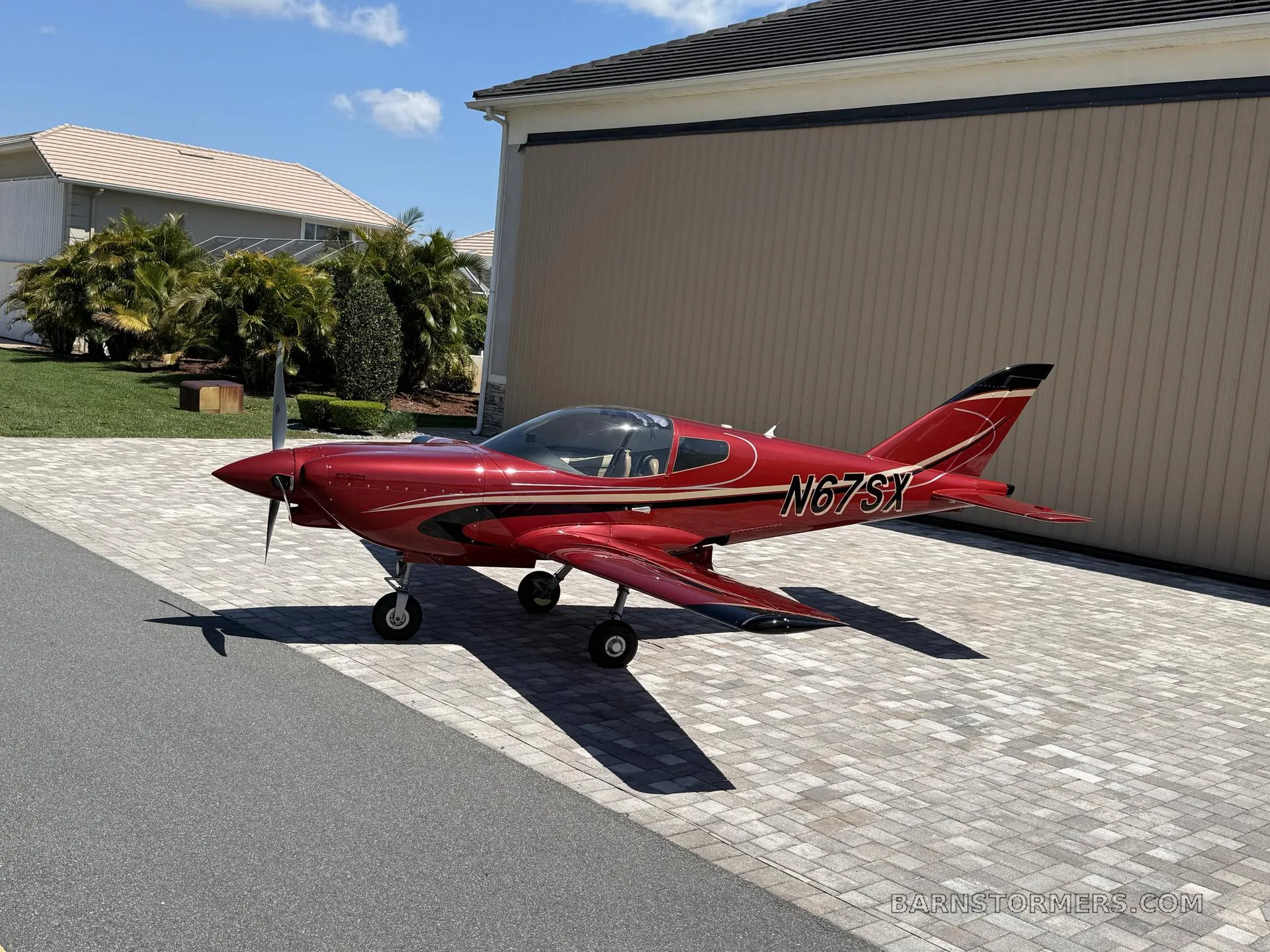 A shiny red single-engine aircraft with registration N67SX is parked on a brick driveway next to a beige house on a sunny day.