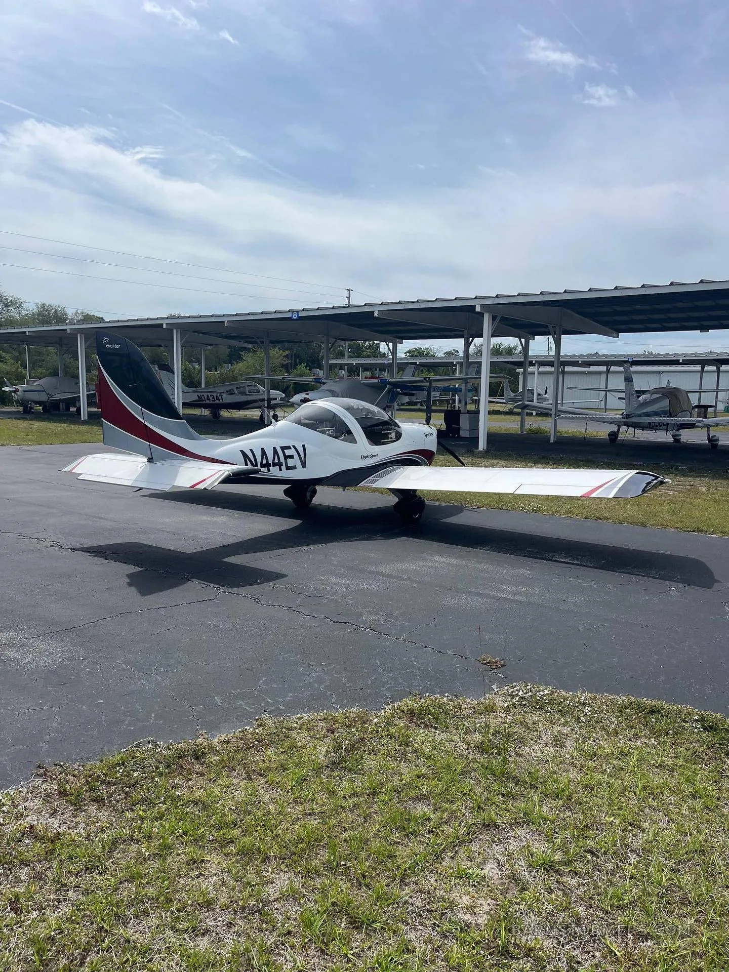 A white and red SportStar aircraft, tail number N44EV, parked on an asphalt surface next to open hangars.