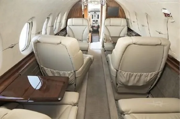 A view down the aisle of a private jet cabin, showing light leather seats and a wooden table, with the cockpit visible in the distance.