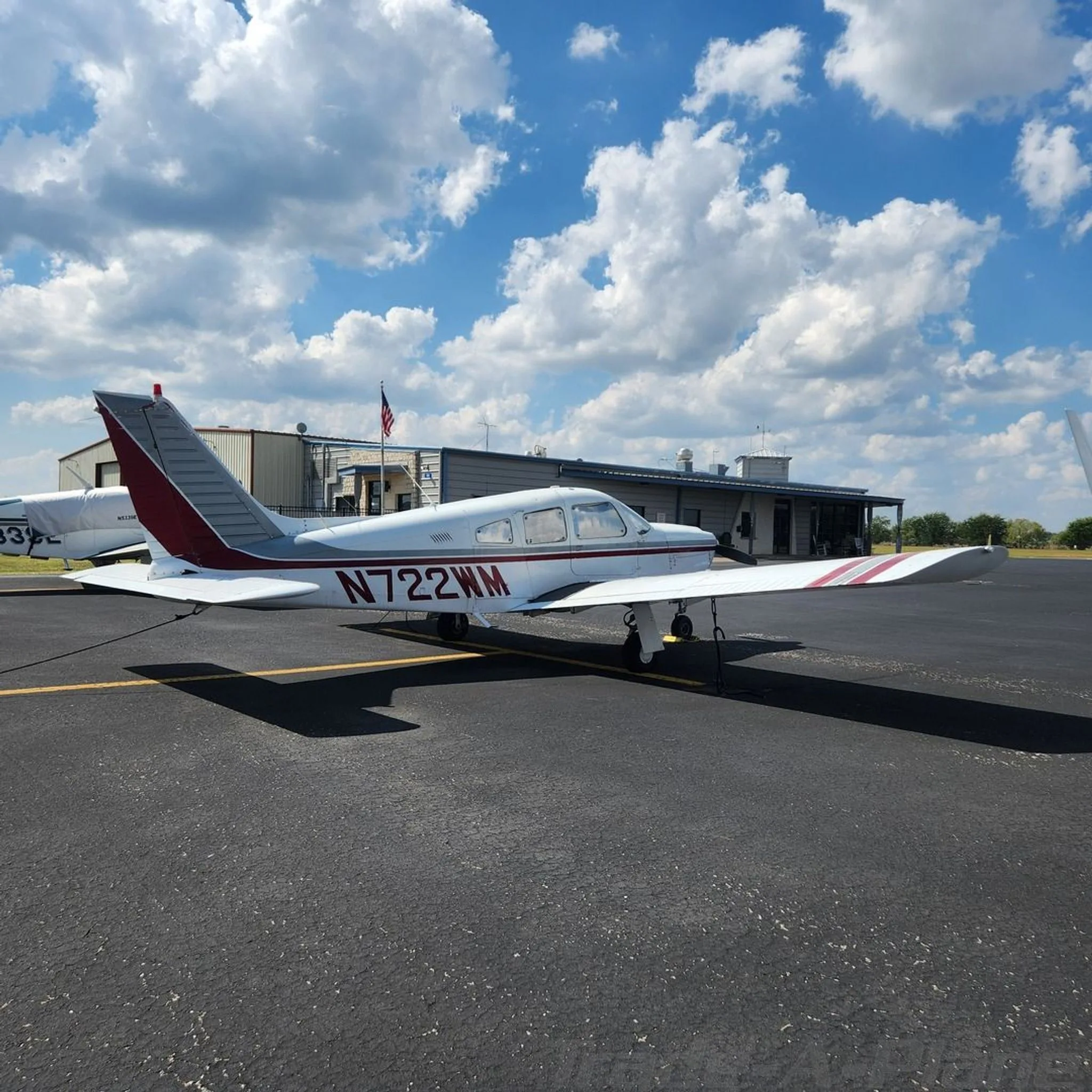 A white Piper Cherokee with red and grey stripes, registration N722WM, parked on an asphalt ramp with a building in the background.