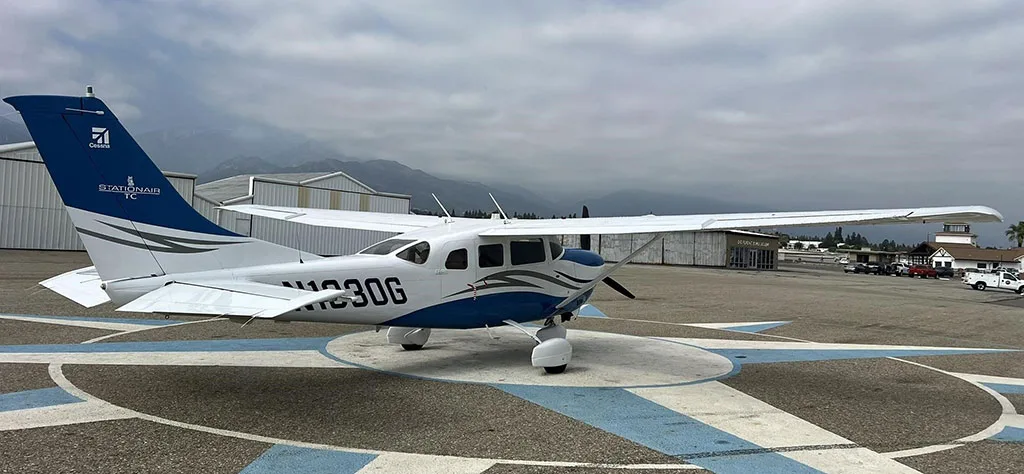 Rear-left view of a white and blue Cessna Stationair TC (N1330G) parked on an airfield with mountains in the background.