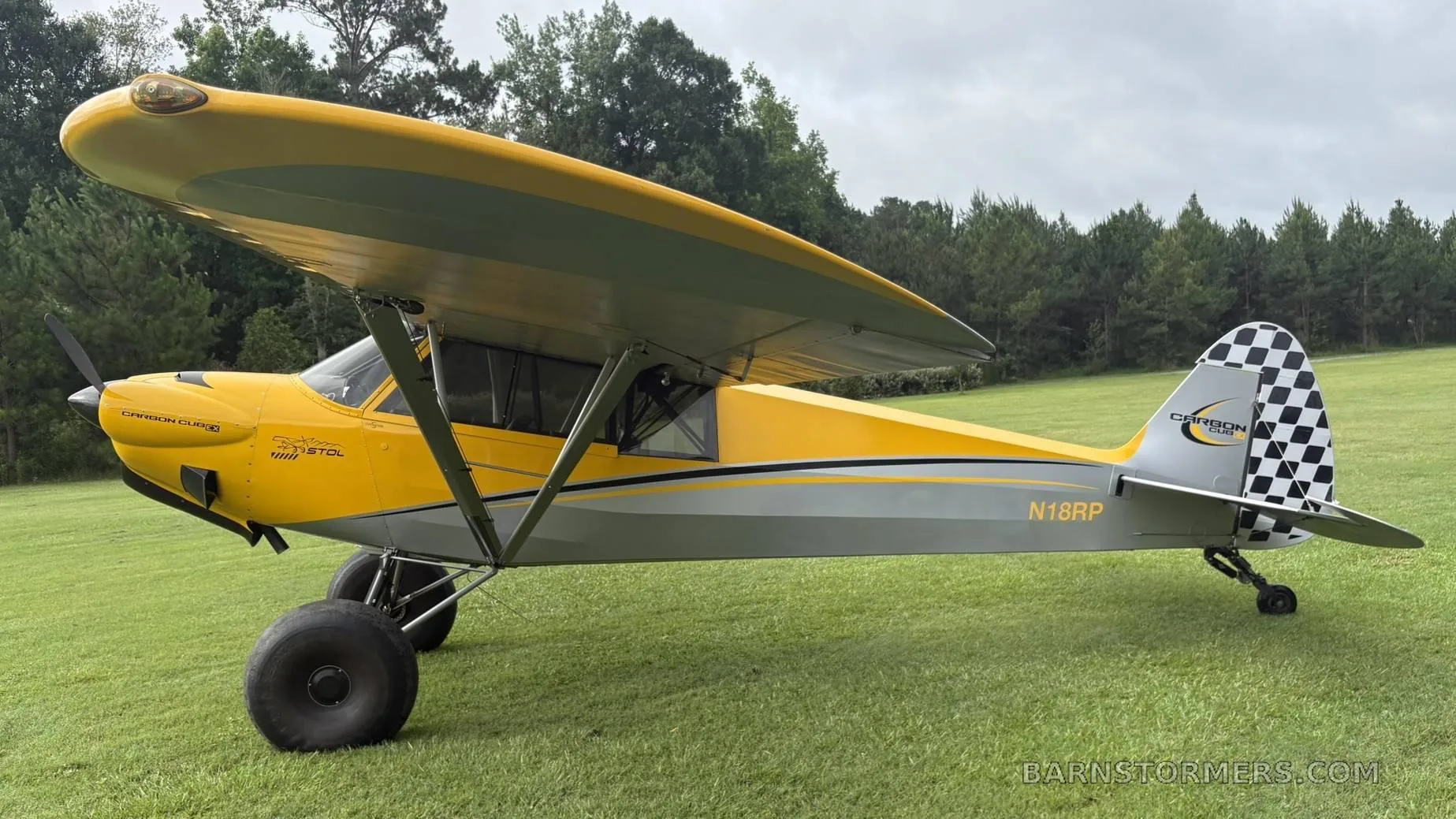 A side view of a yellow and grey CubCrafters Carbon Cub aircraft with registration N18RP and a checkered tail, parked on a grassy field.