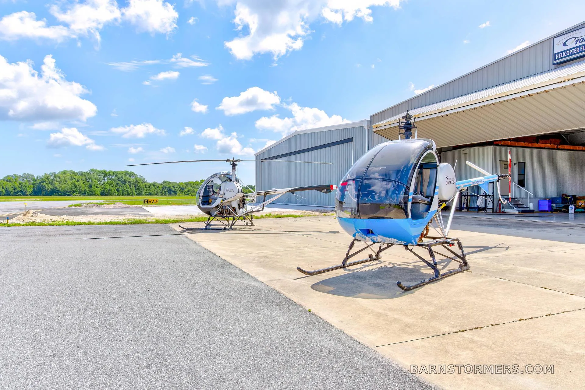 Two Schweizer 300CBi helicopters, one blue and one white, are parked on an airfield apron in front of a hangar.