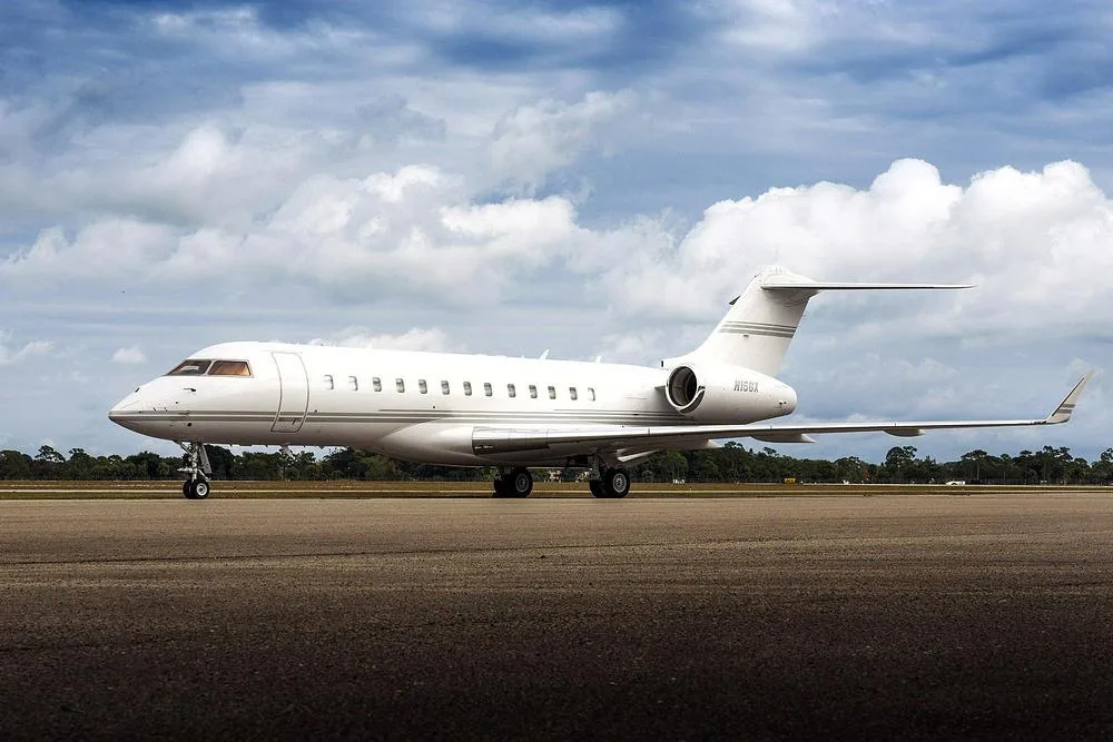 A white Bombardier Global Express business jet, registration N15GX, parked on a tarmac with trees in the background.