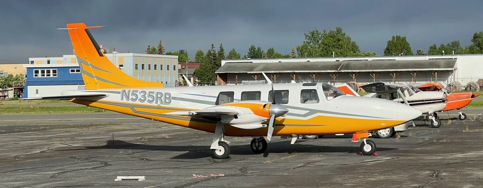 A white and orange twin-engine Piper Navajo aircraft (N535RB) parked on an asphalt ramp with other aircraft and buildings in the background.