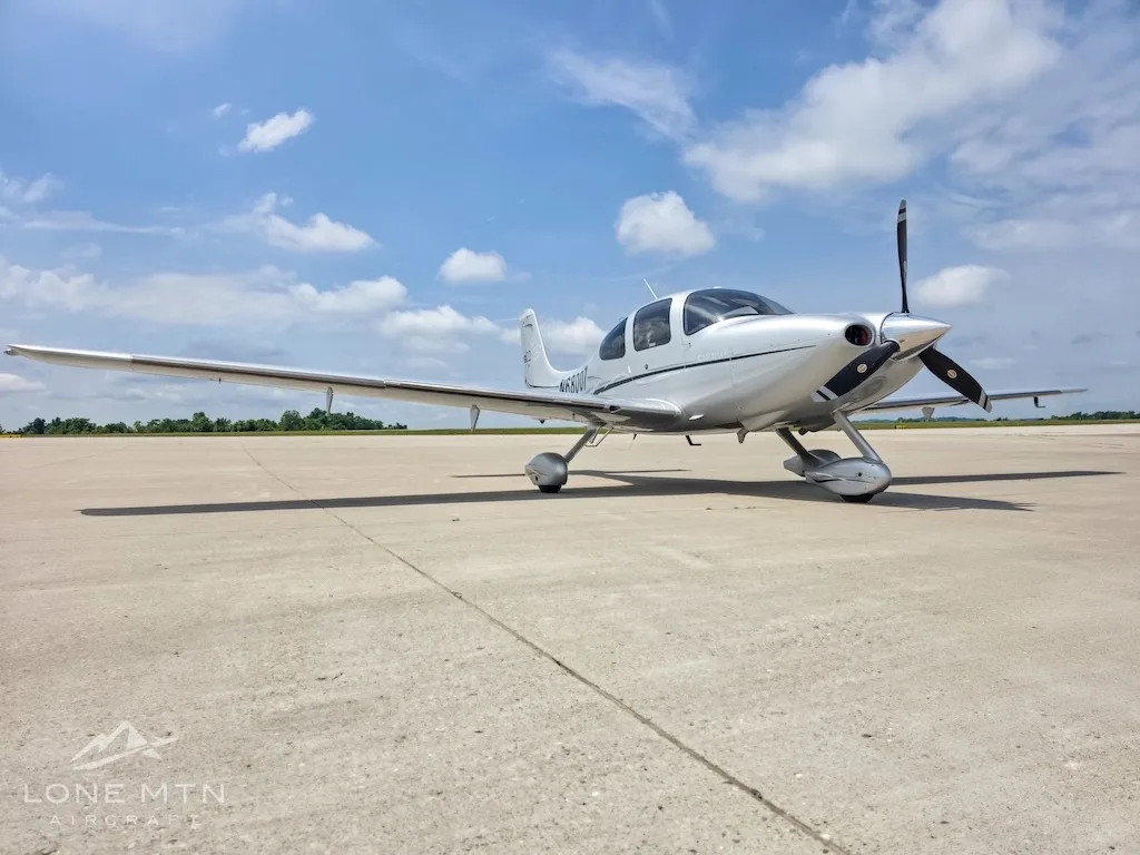 A silver Cirrus SR22 aircraft (N68007) with a black propeller, parked on an airfield under a partly cloudy sky, viewed from the front-left.