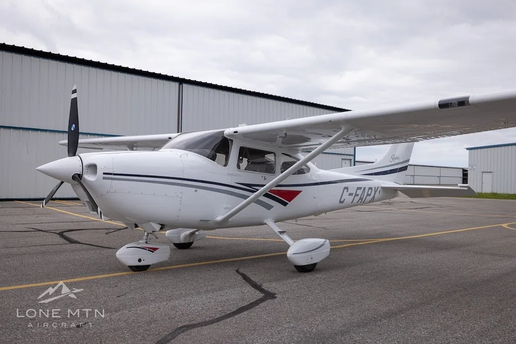 A white Cessna 182 Skylane with grey and blue stripes, registration C-FAPX, parked on an asphalt ramp in front of hangars.