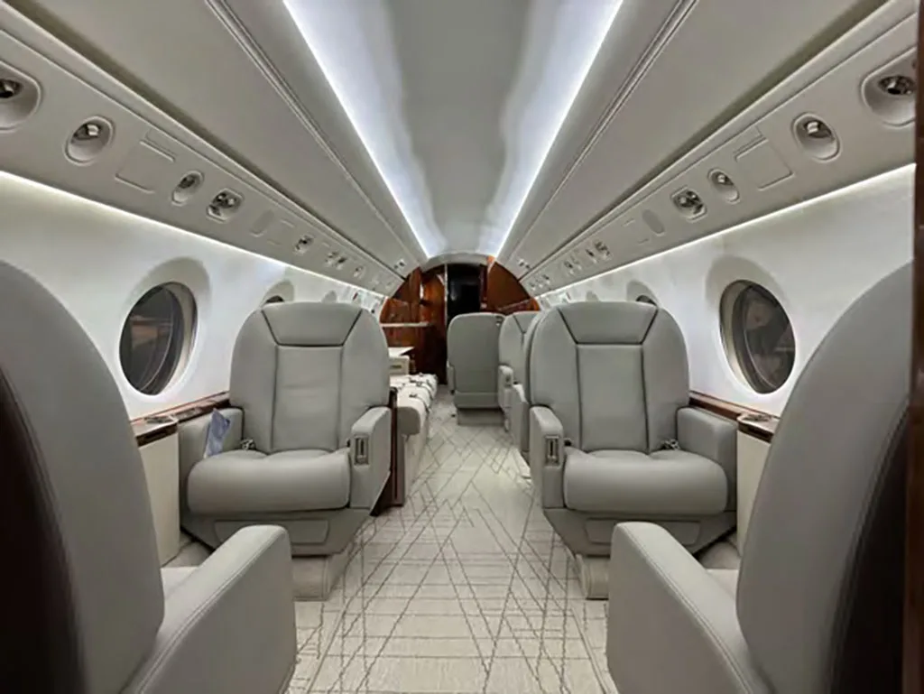 A long view down the aircraft cabin aisle, showing rows of light gray leather seats and a patterned carpet.