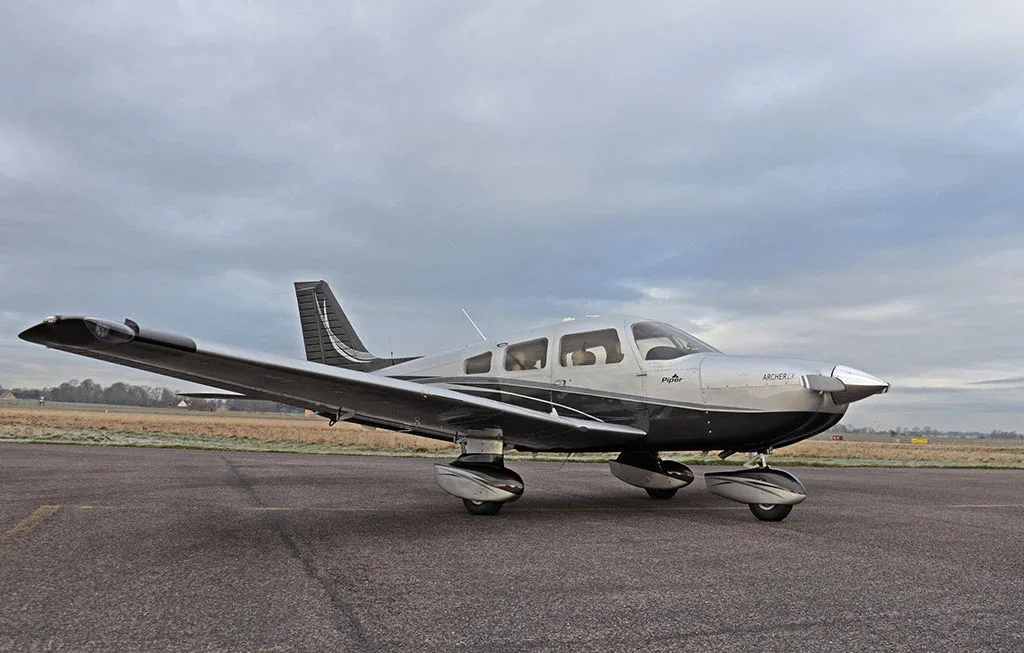 A side view of a silver and black Piper Archer LX aircraft on an airfield with a cloudy sky.