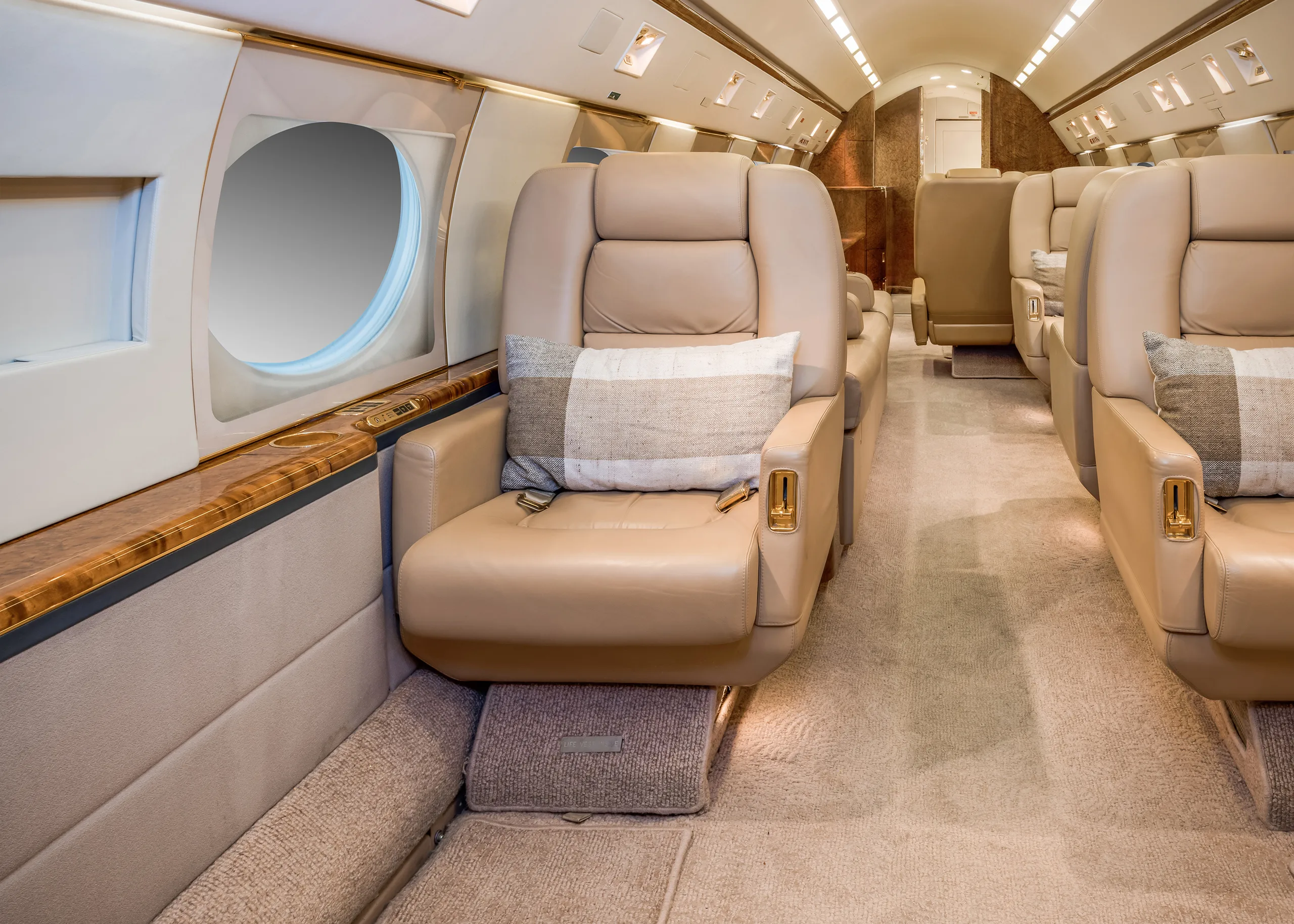 A close-up view of a beige leather aircraft seat with a plaid pillow, next to a large oval window.