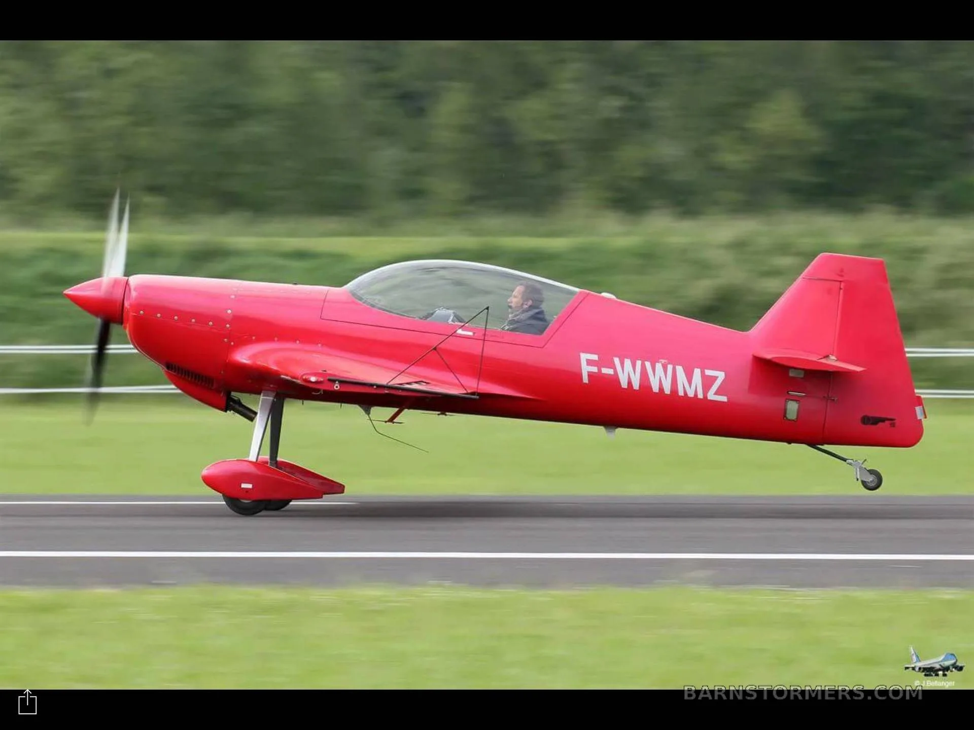 A red single-engine aerobatic aircraft with registration F-WWMZ is taking off from a runway, with a pilot visible in the cockpit.