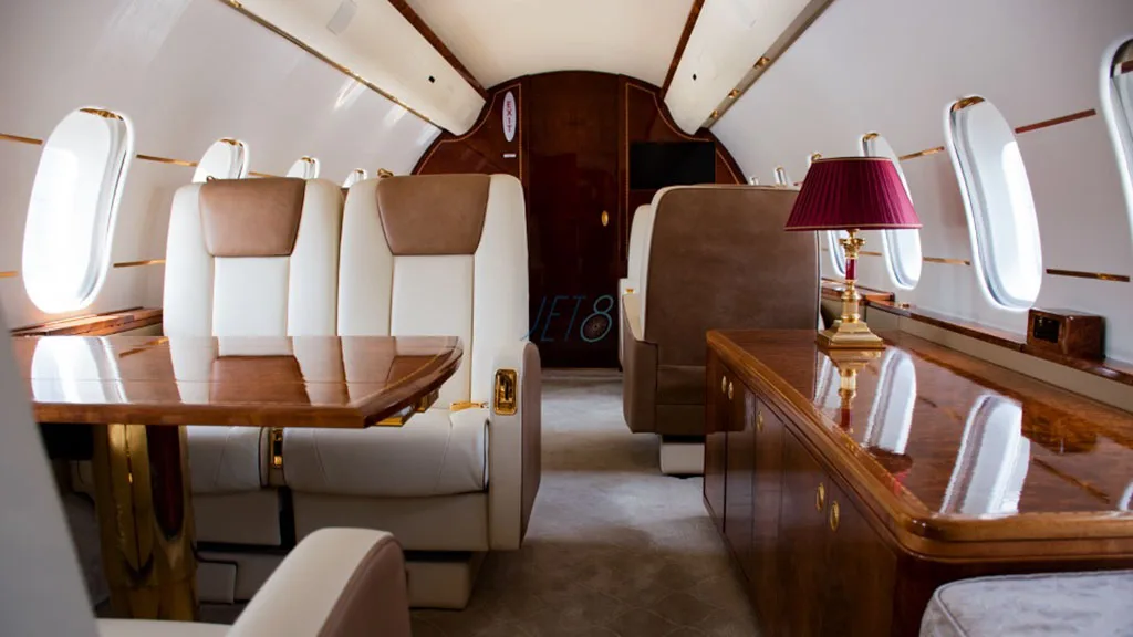 A luxurious aircraft cabin featuring a polished wooden table, cream and brown leather seats, and a red-shaded lamp.