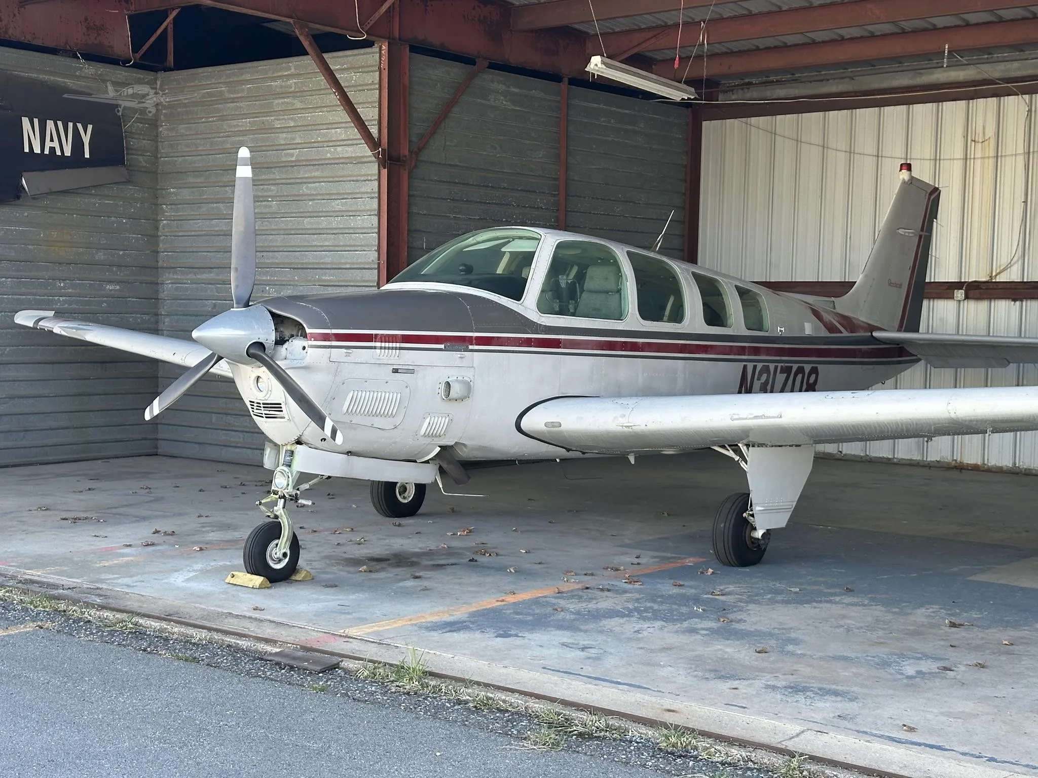 A silver and maroon single-engine propeller aircraft, likely a Beechcraft Bonanza with tail number N31708, parked inside a hangar.