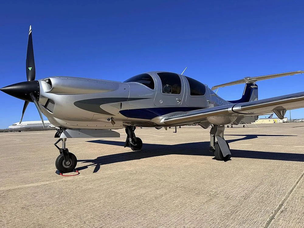 A side view of the silver and blue Diamond DA40 aircraft, N66RG, parked on a sunny tarmac with other planes in the distance.