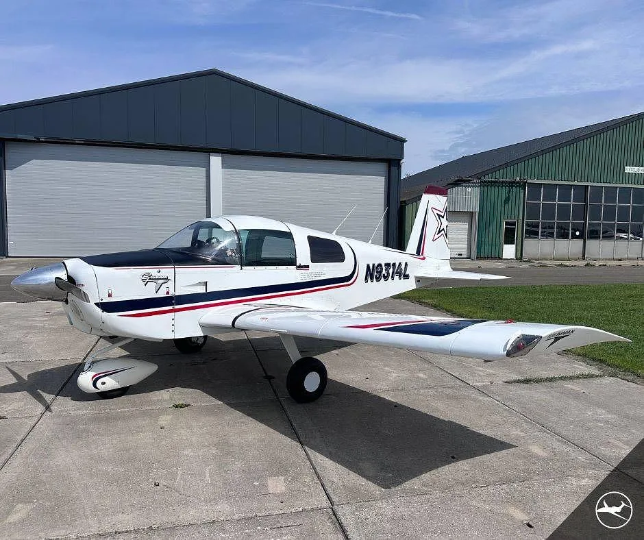 A white Grumman AA-5B Tiger aircraft with black and red stripes, registration N9314L, parked on concrete in front of hangars.