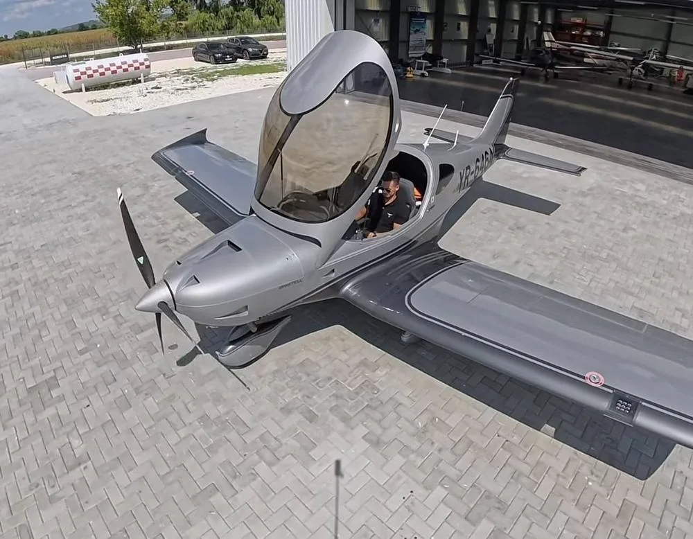 An overhead view of a silver Pipistrel Virus SW aircraft (YR-6464) with its canopy open and a person in the cockpit, parked outside a hangar.