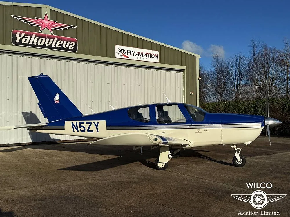 A blue and white single-engine aircraft, N5ZY, parked on an airfield in front of a hangar with 'Yakovlev' and 'Fly Aviation' signs and bare trees in the background.