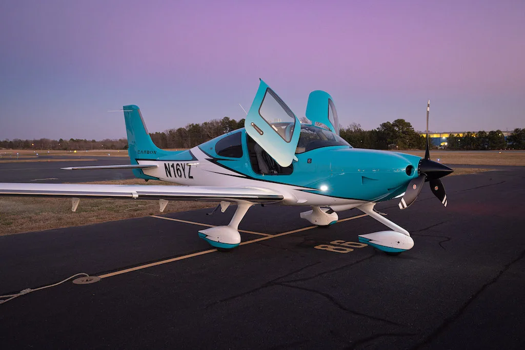 A turquoise and white Cirrus SR22 aircraft with its doors open, parked on an airfield at dusk.