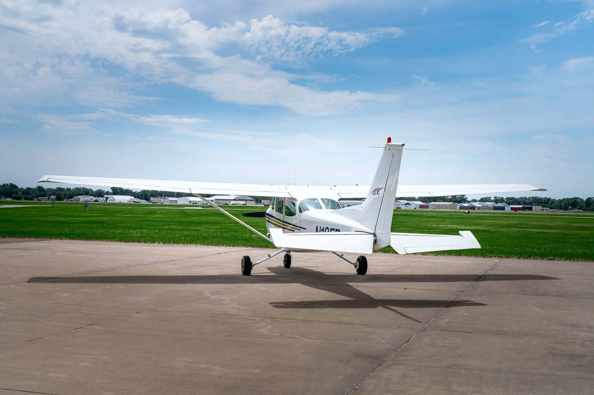 A white Cessna 172 with blue, gold, and red stripes on the fuselage, parked on an asphalt ramp, viewed from the rear.