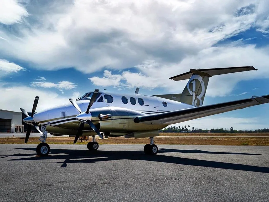 A silver and gold twin-turboprop aircraft with a large '1B' logo on its tail, parked on an asphalt tarmac under a partly cloudy sky.