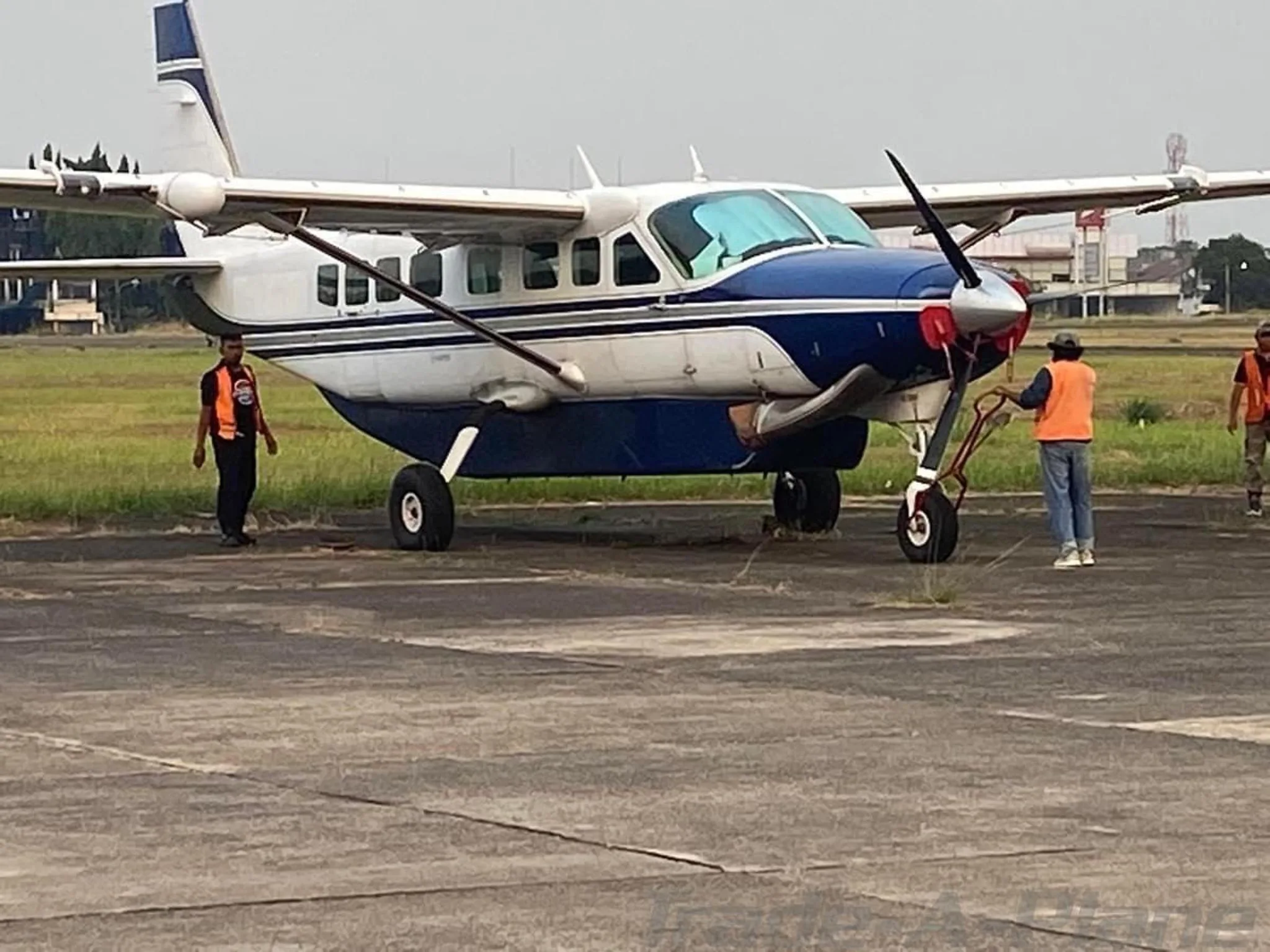 A white and blue single-engine turboprop aircraft, likely a Cessna Caravan, on an airfield with ground crew.